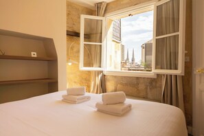 A hotel room with a bed, two towels, a window with curtains, and a view of buildings.