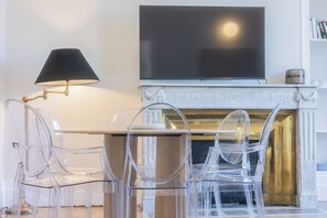 A modern living room with a flat-screen TV mounted above a fireplace, clear plastic chairs, and a floor lamp.
