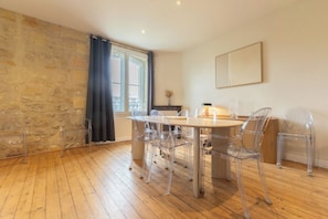 Un área de comedor con una mesa redonda y sillas transparentes, una pared de piedra, una ventana con cortinas y piso de madera.