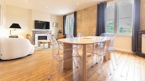 Un moderno salón con suelo de madera, una mesa de comedor redonda con sillas transparentes, una chimenea y ventanas grandes con cortinas.