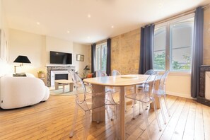 A modern living room with a wooden floor, a round dining table with clear chairs, a fireplace, and large windows with curtains.