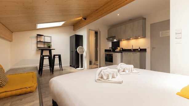 A bedroom with a bed, a desk, a chair, a fan, and a kitchenette.