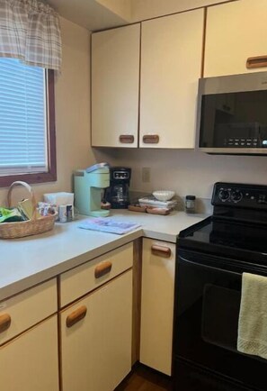 Cocina pequeña con microondas, estufa y cafetera. Hay una ventana con persianas y una cesta en la encimera.