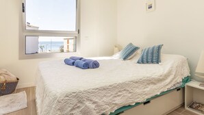 A bedroom with a large window, a bed with white bedding and blue pillows, a bedside table with a lamp, and a view of the sea. - El Fenua by Interhome (St Antoni de Calonge)