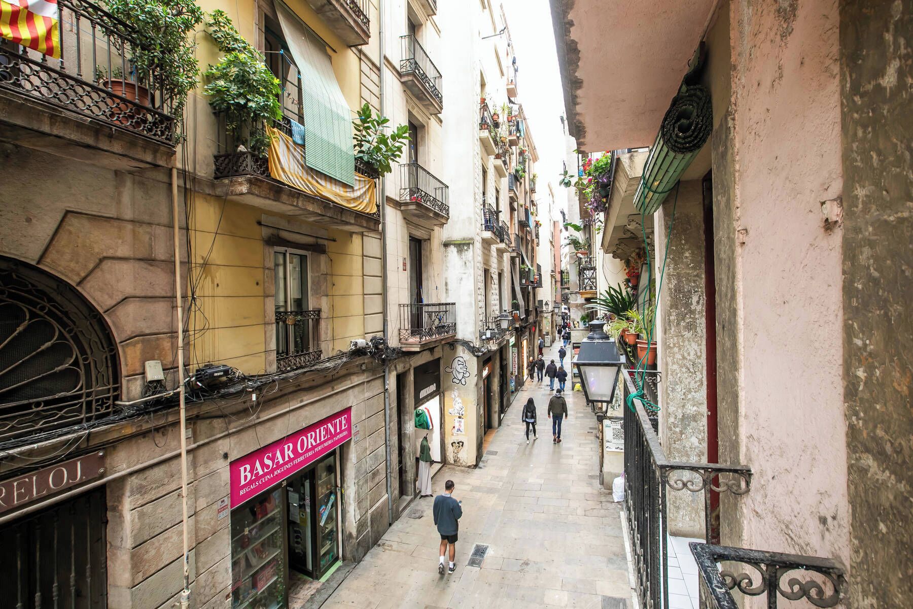 Un carrer urbà estret amb edificis a ambdós costats, una botiga amb cartell i vianants caminant.