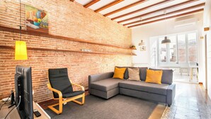 A living room with a brick wall, a grey sofa with yellow cushions, a wooden chair, a television, and a large window.