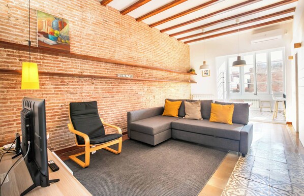 Una sala de estar con pared de ladrillo, un sofá gris con cojines amarillos, una silla de madera, un televisor y un ventanal grande.
