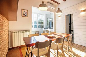 Un coin repas avec une longue table et des chaises, un radiateur et une fenêtre donnant sur un espace verdoyant.