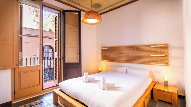 A bedroom with a bed, bedside tables, a hanging light fixture, and a view through an open door.