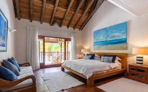 Un dormitorio con cama de madera, ropa de cama blanca, vistas al exterior, un cuadro de una playa y techo de madera.