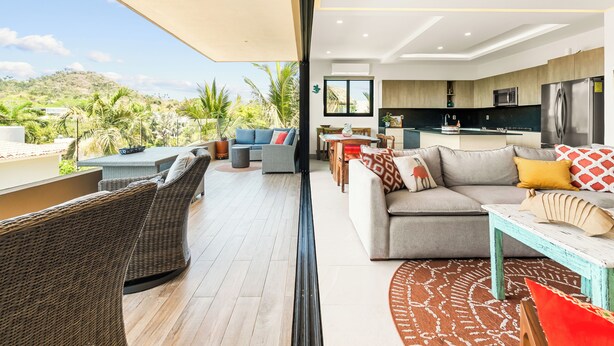 A modern living area with a balcony, furnished with wicker chairs and a sofa, a dining table, and a kitchen in the background. - Manta Morada: Mar, San Pancho, Beachfront and Ocean views (San Francisco)