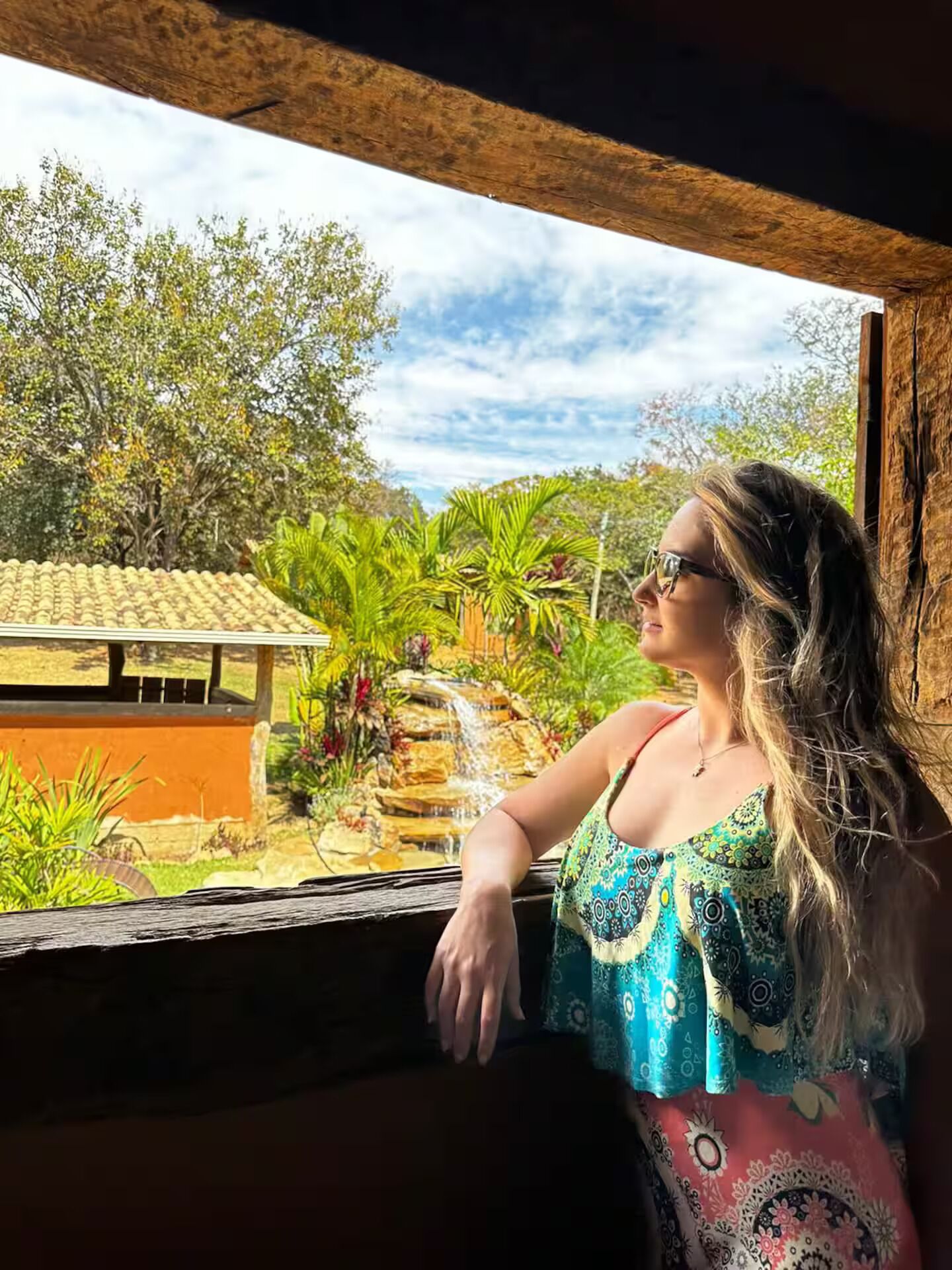 A person leaning on a window with a view of a garden and a building with a tiled roof.