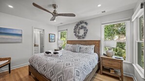 A bedroom with a large bed, a ceiling fan, a wooden headboard, a nightstand with a lamp, and a window with a view of greenery. - Modern Beach Home on Quiet Street w/Private Pool. 1 Block to Beach! (Isle Of Palms)