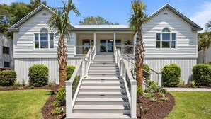 A two-story house with a front porch, a staircase leading to the entrance, and a well-maintained lawn with palm trees. - Modern Beach Home on Quiet Street w/Private Pool. 1 Block to Beach! (Isle Of Palms)