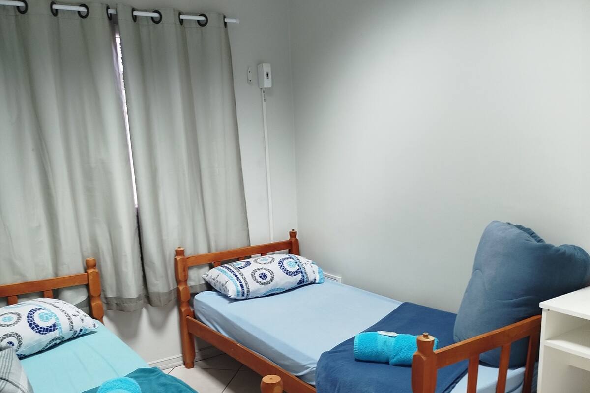 A small, tidy room with two wooden beds, each with blue bedding and pillows. There is a white desk with a chair and a small trash bin.