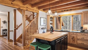 A kitchen with a wooden island, green bar stools, a wooden staircase, and large windows with a forest view.