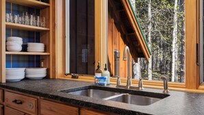 A kitchen with wooden cabinets, a granite countertop, a double sink, and a window overlooking a forest.