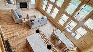 Geräumiges Ferienhaus-Interieur mit Holzboden, Kamin aus Stein, einer Couch, einem Esstisch mit Stühlen und großen Fenstern mit Blick ins Freie.