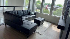 Een moderne woonkamer met een hoekbank, een klein voetenbankje en grote ramen met uitzicht op de omgeving.