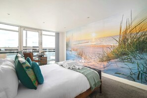Een slaapkamer met een groot raam, een bed met kussens, een schilderij van een strand en uitzicht op het water.