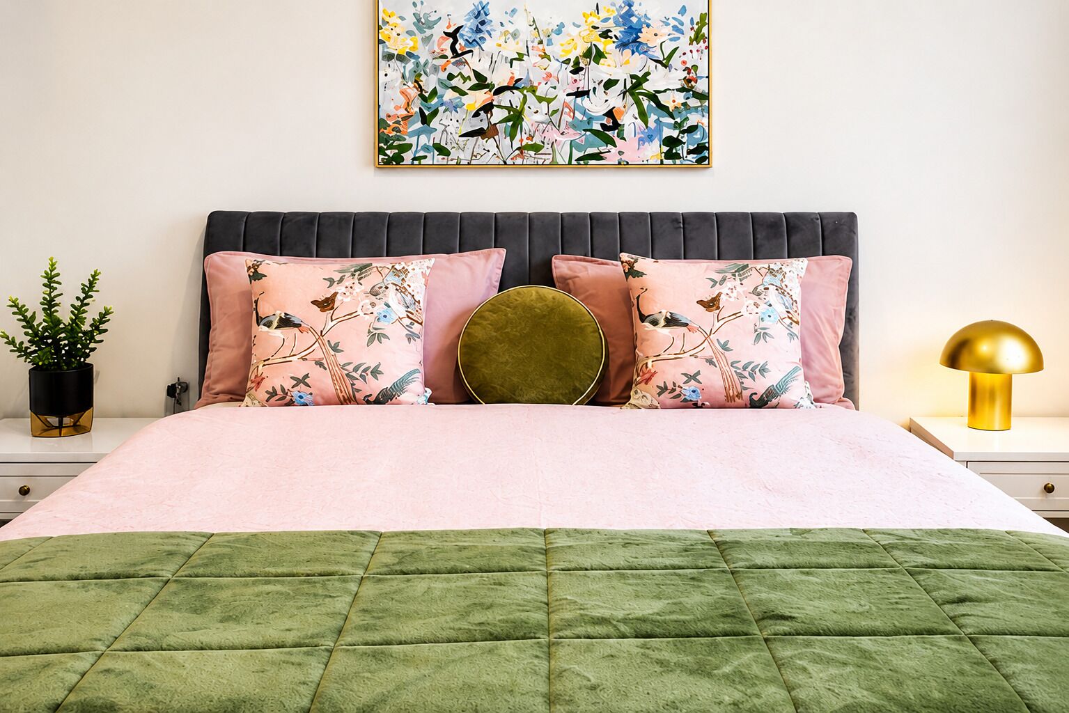 Un lit recouvert d’une couette verte, agrémenté de deux coussins à motifs floraux, d’un coussin rond doré servant d’élément décoratif, et d’un tableau abstrait accroché au mur.