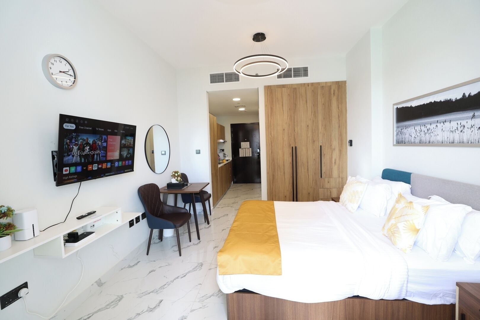 A hotel room with a bed, a desk with a chair, a TV mounted on the wall, a wardrobe, and a painting on the wall.