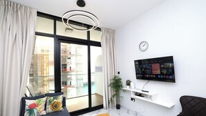A modern living room with a large window, a flat-screen TV, a sofa with floral cushions, a small plant, and a clock on the wall.