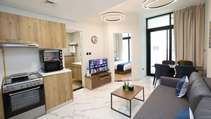 A modern hotel room with a kitchenette, a living area with a sofa and coffee table, a TV, and a large window with curtains.