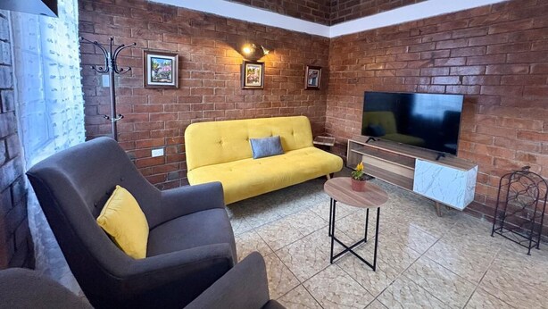 A cozy living room with a brick wall, a yellow sofa, a grey armchair, a small table, and a TV.