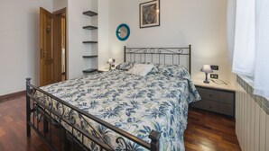 Un dormitorio con cama, mesitas de noche, estante en la pared, una puerta y una ventana con cortinas.