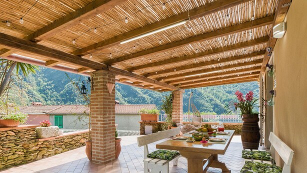 Un patio al aire libre con una pérgola de madera, paredes de piedra y una mesa preparada para comer.