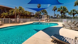 A swimming pool with a blue shade sail, palm trees, and a fenced area with lounge chairs. - Shared Pool, Pet Friendly, Garage Access (Port Aransas)