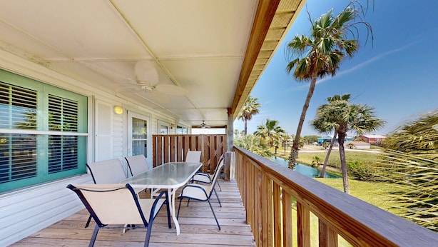 A covered outdoor patio with a table and chairs, wooden railings, and palm trees in the background. - Shared Pool, Pet Friendly, Garage Access (Port Aransas)