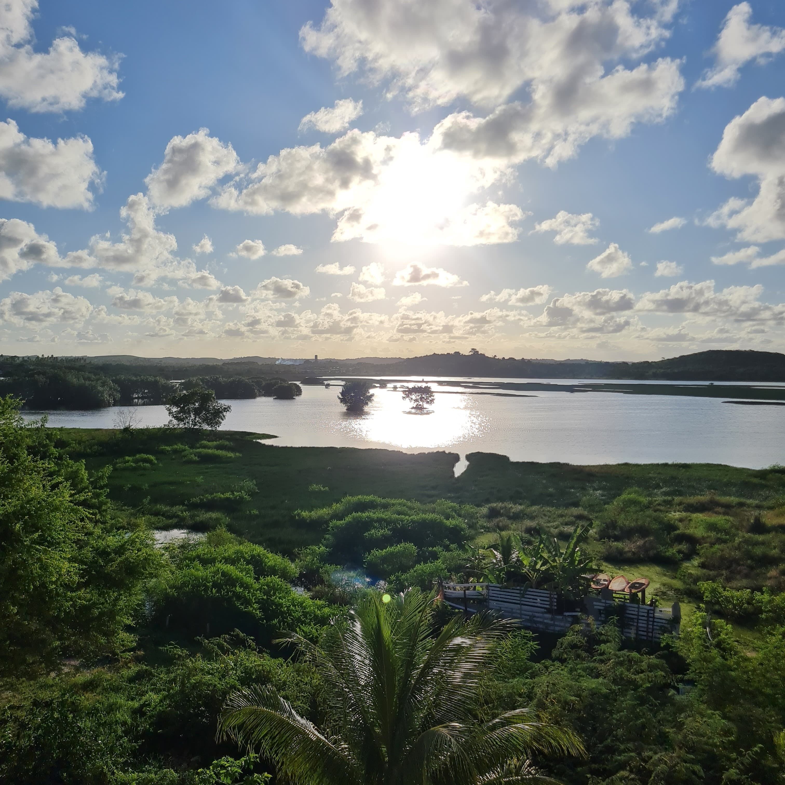 A view of a lake surrounded by lush greenery and a clear sky with scattered clouds.