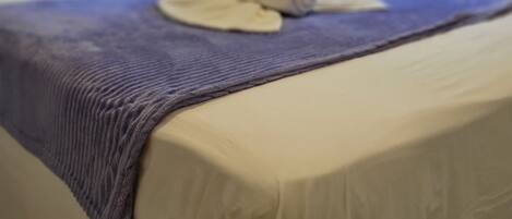 A neatly made bed with a blue blanket and rolled towels, a bedside table, and a window with a view of greenery.