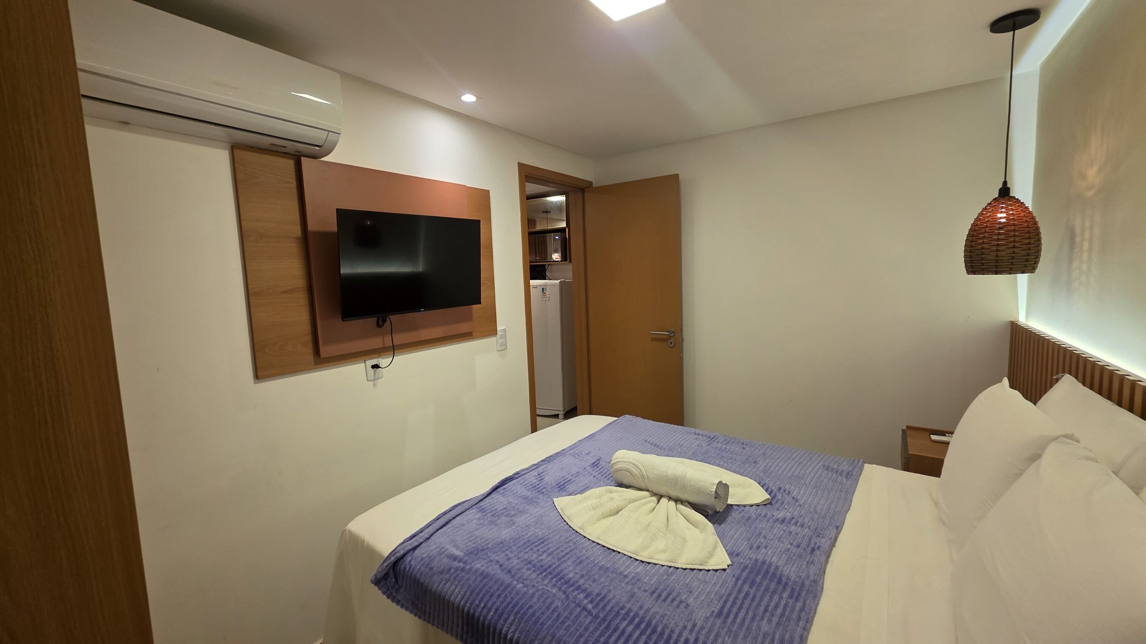 A hotel room with a bed, a television mounted on the wall, an air conditioning unit, and a door leading to a bathroom.