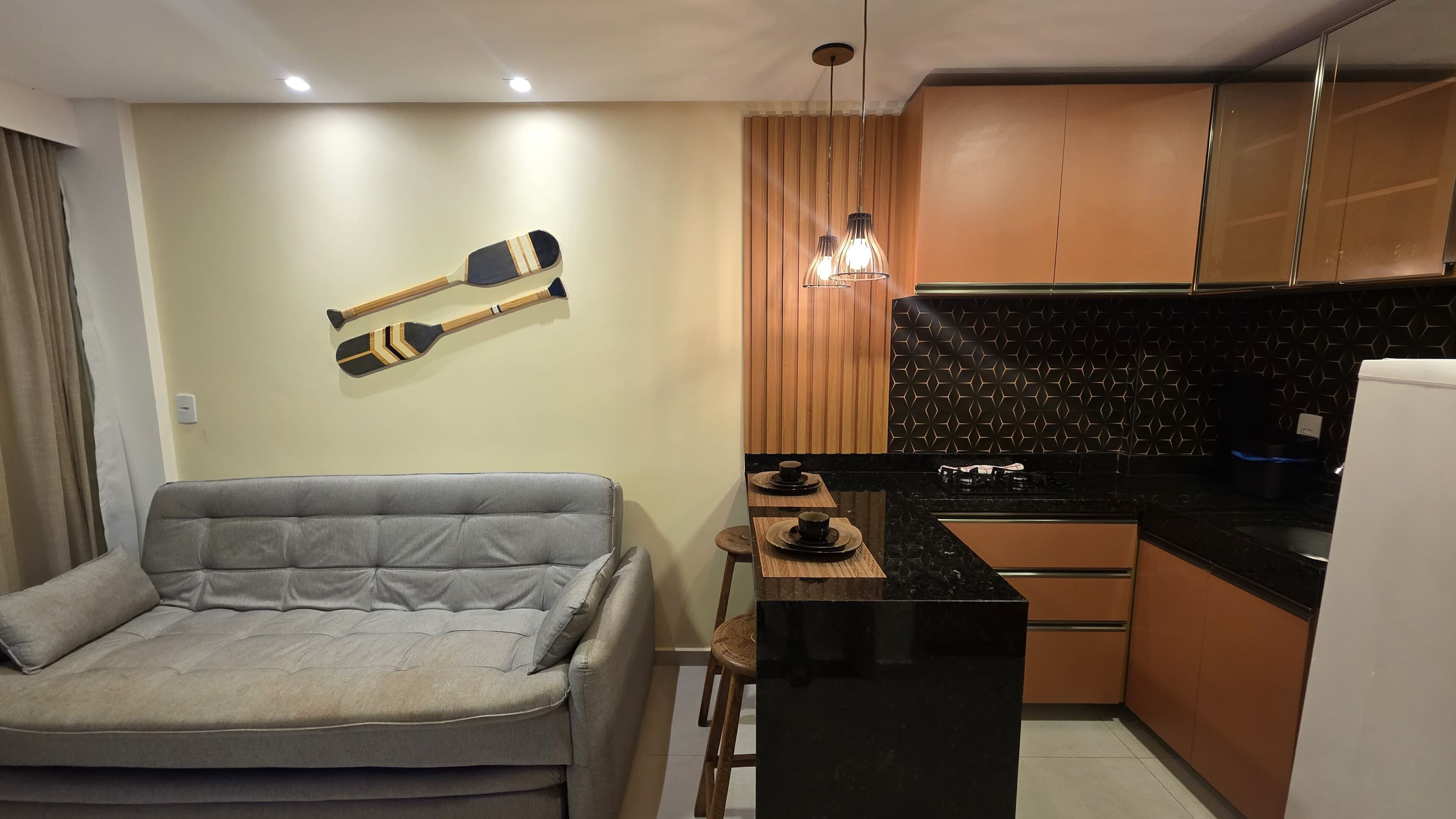 A compact living space with a grey sofa, a small dining table with two plates, and a kitchen area with wooden cabinets and a black countertop.