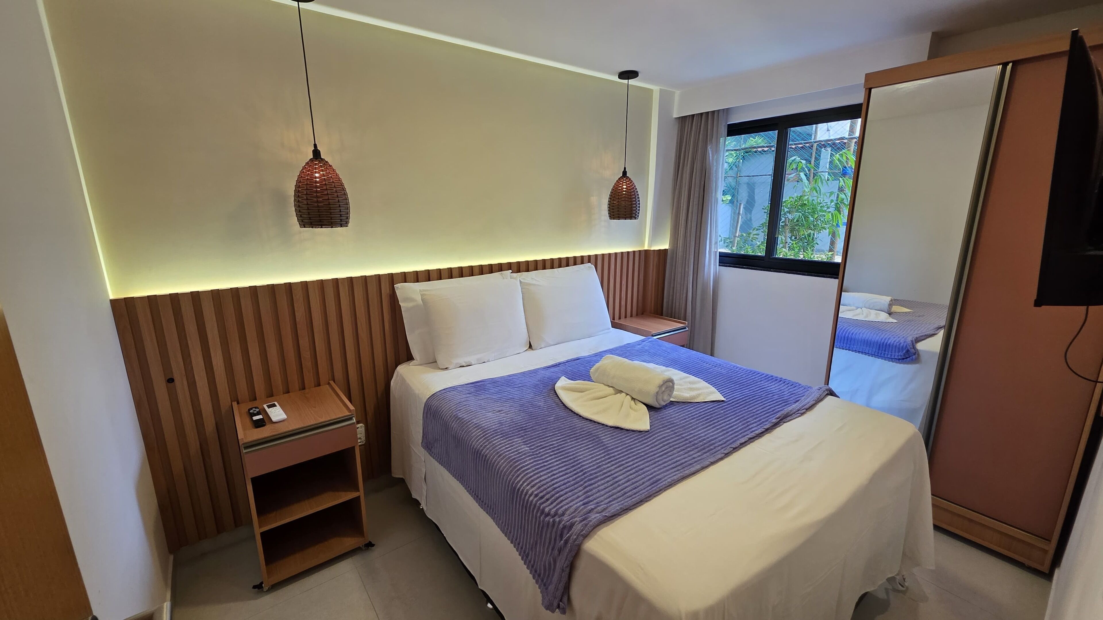 A hotel room with a bed, bedside table, and a window with a view of greenery.