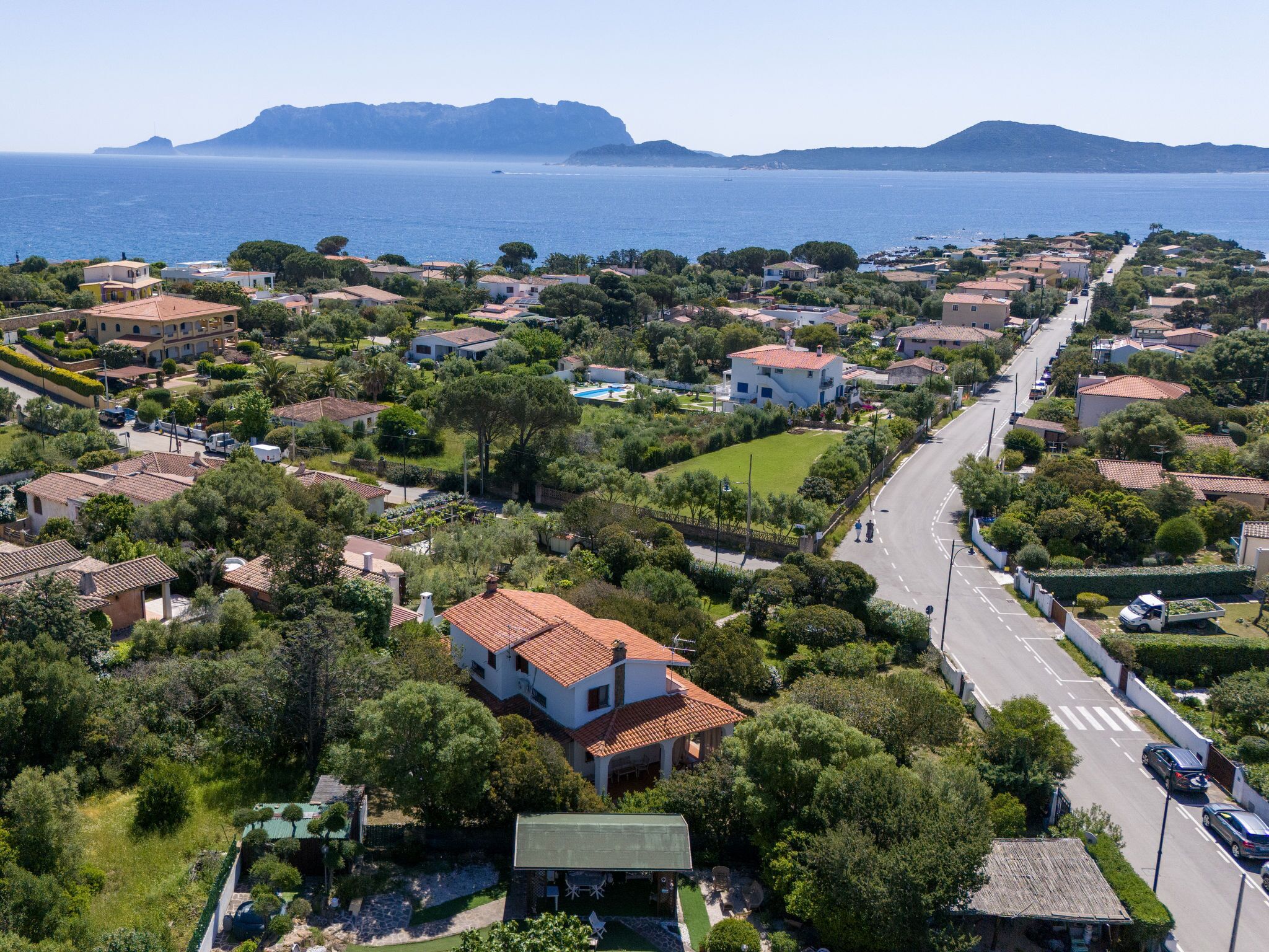 Ett flygbildsbild av ett bostadsområde vid kusten med hus, en väg och en utsikt över havet och bergen i bakgrunden.