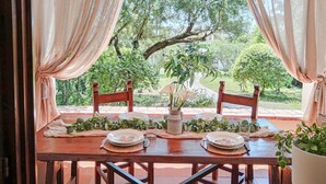 A dining table set with plates, glasses, and a vase of flowers, surrounded by curtains and chairs, with a view of greenery outside. - Casa Tonina by Interhome (Pittulongu)