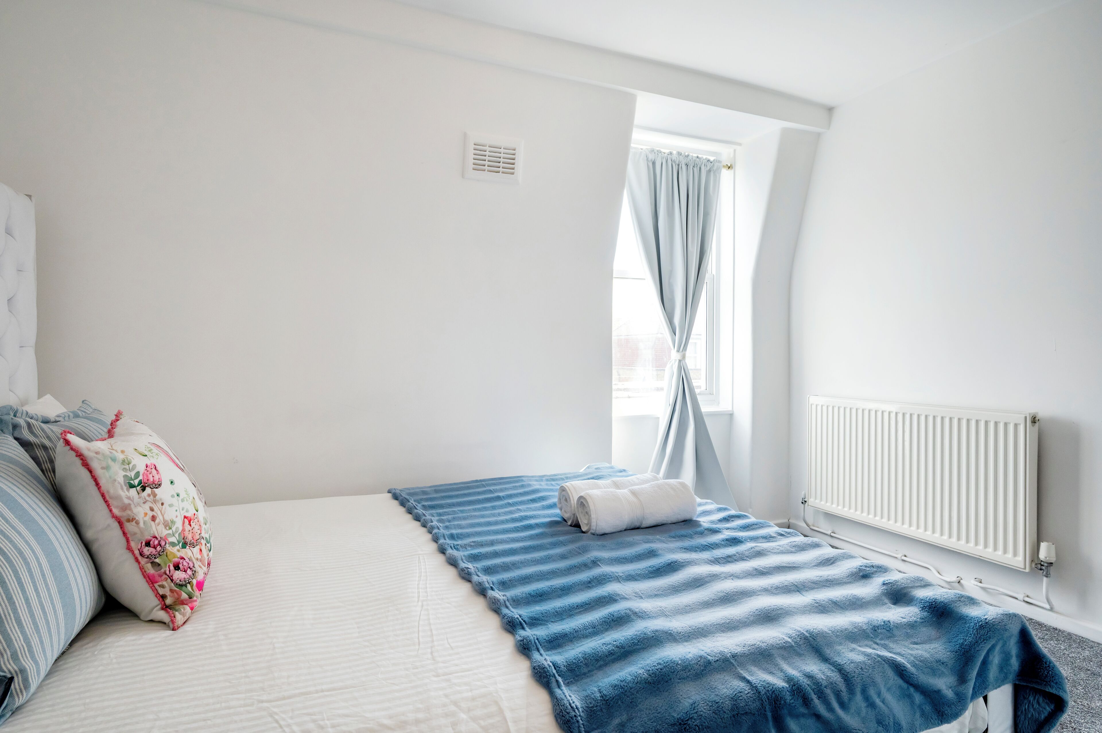 Ein Schlafzimmer mit Bett, einem Fenster mit Vorhängen, einem Heizkörper und einem Wandlüfter.