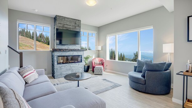 A modern living room with a stone fireplace, a grey sofa, a blue armchair, and large windows offering a view of the outdoors. - 2 Side-by-Side Ski-In/Ski-Out Townhomes | Sleeps 19 w/ Views (Silver Star Mountain)