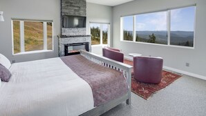 A bedroom with a large bed, a stone fireplace, and a view of a forest through large windows. - 2 Side-by-Side Ski-In/Ski-Out Townhomes | Sleeps 19 w/ Views (Silver Star Mountain)