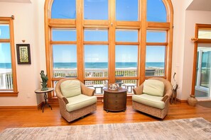 A room with a large bay window overlooking the beach, wicker chairs, a small side table, and a decorative vase.