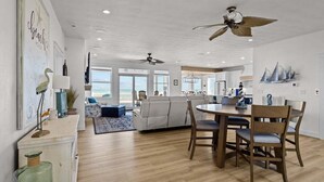 A spacious living area with a large window overlooking the ocean, a dining table with chairs, and a ceiling fan. - A Wave From It All | Oceanfront | Hot Tub (Virginia Beach)