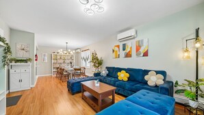 A modern living room with a blue sofa, wooden coffee table, and a dining area with a white cabinet and a plant. - Spacious Modern 3BR Free Parking 30 min to NYC (Jersey City)