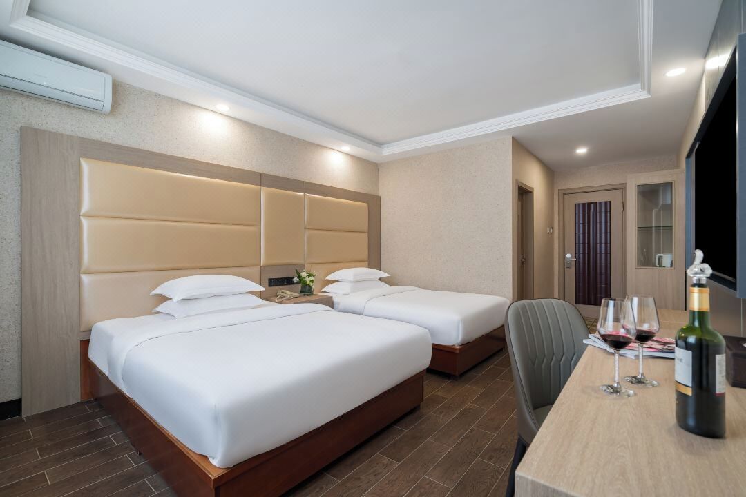 Habitación de hotel con dos camas, cabecero de madera, mesita de noche con lámpara, y una mesa con copas de vino y una botella.