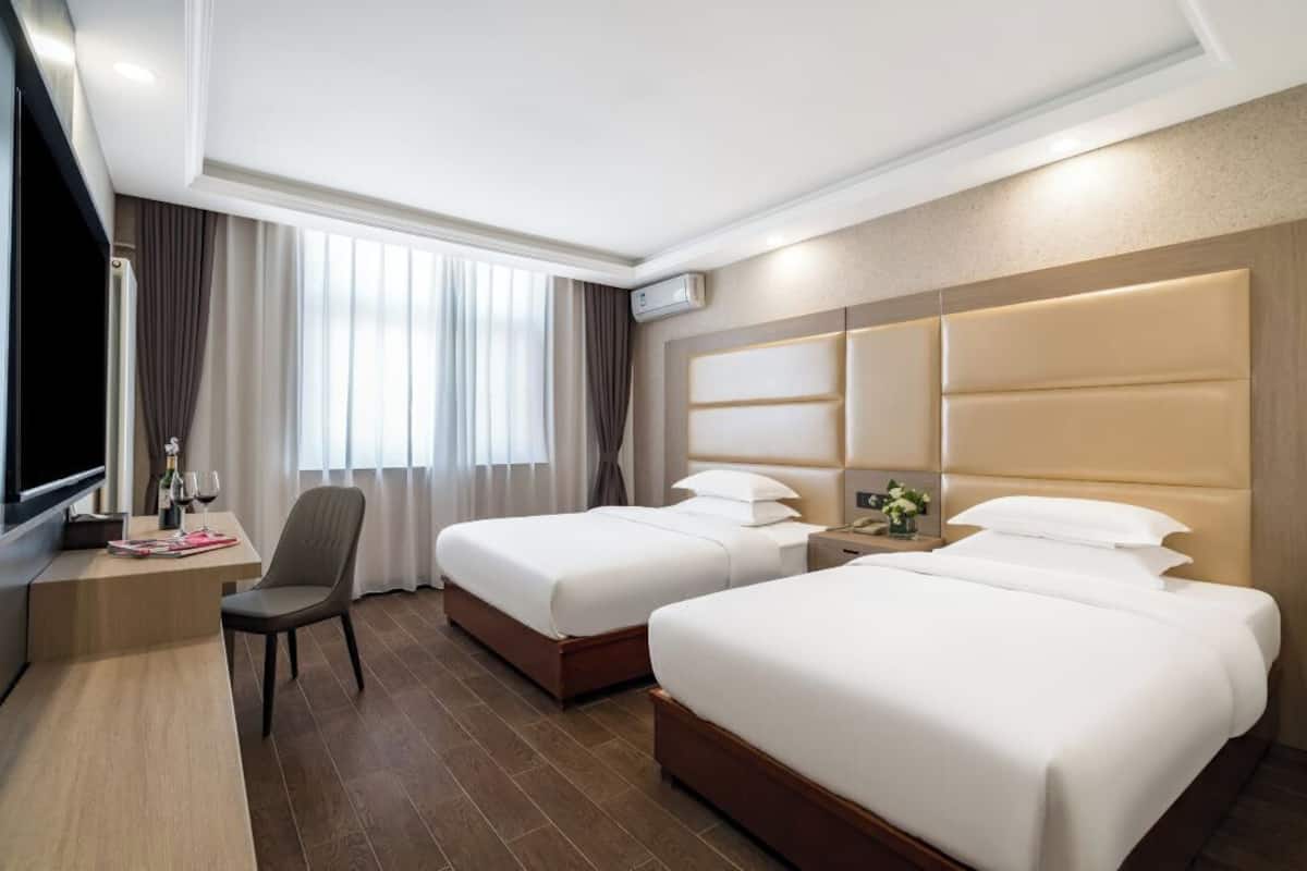 A hotel room with two beds, a desk with a chair, a TV, and a large window with curtains.