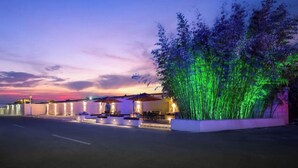 Een modern buitenterrein met tafels en parasols, omgeven door een aangelegde tuin met verlichte bamboe.