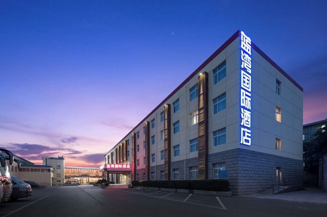 A modern hotel building with a prominent sign, multiple floors, and a parking lot in the foreground.